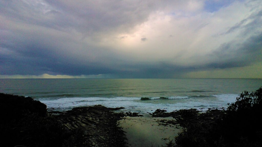 Paysage océanique contrasté entre rochers sombres eau turquoise et ciel lavande chargé de nuages, avec au loin une éclaircie